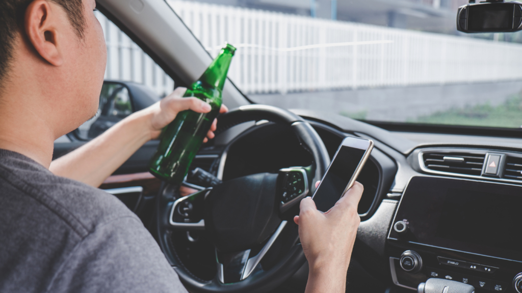 Image of a man driving with a beer in his hand and a cell phone in the other